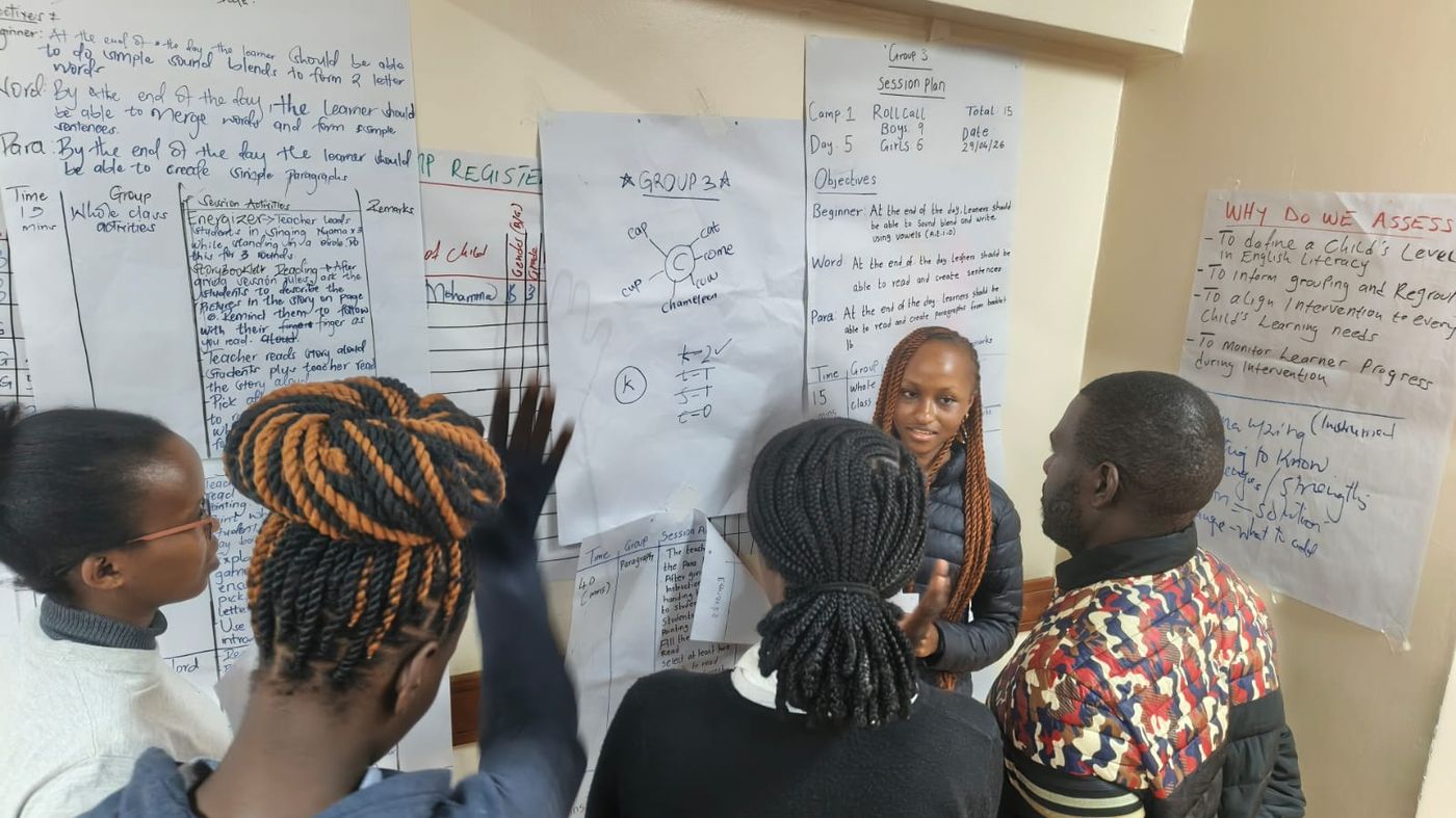Team members discussing a poster titled 'Why Do We Assess' outlining the M&E rationale